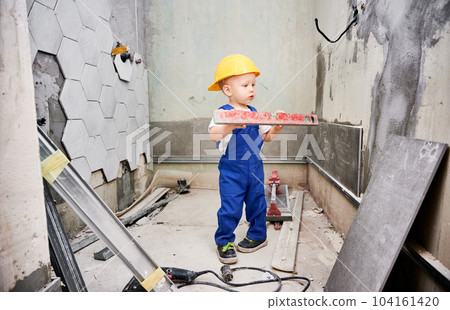 Boy construction worker holding spirit level instrument. Kid in safety helmet and work overalls standing in apartment bathroom. Home renovation and childhood concept. Boy construction worker holding spirit level instrument. Kid in safety helmet and work overalls standing in apartment bathroom. Home renovation and childhood concept. 104161420