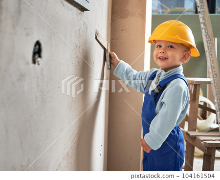 Happy little boy builder worker looking away and smiling while measuring wall with construction ruler. Cheerful kid in safety helmet and work overalls working on home renovation. 104161504