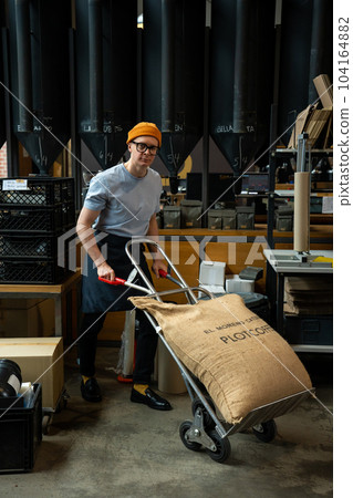 Roastery worker standing with sacks with coffee beans uploading. Roastery worker standing with sacks with coffee beans uploading. 104164882