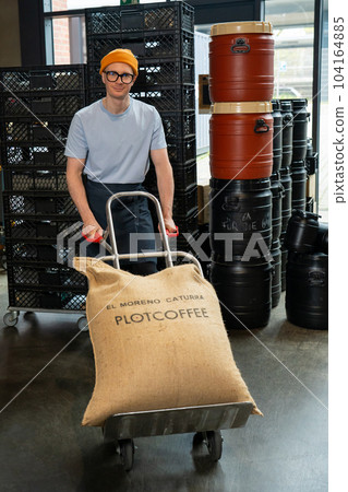 Roastery worker standing with sacks with coffee beans uploading. Roastery worker standing with sacks with coffee beans uploading. 104164885