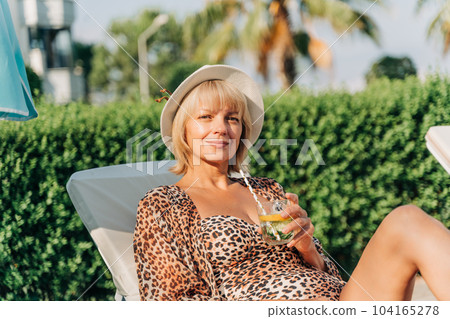 Portrait of happy mature adult woman relaxing deck chair near the pool and looking at camera. Beautiful caucasian senior woman in hat drink lemonade cocktail enjoying summer vacation 104165278