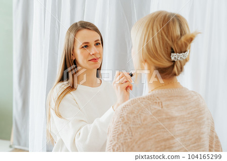 Beautiful smiling young caucasian mature senior mother mom and adult daughter doing makeup, standing near mirror, and looking at reflection in living room home. Family with two women relax together Beautiful smiling young caucasian mature senior mother mom and adult daughter doing makeup, standing near mirror, and looking at reflection in living room home. Family with two women relax together 104165299