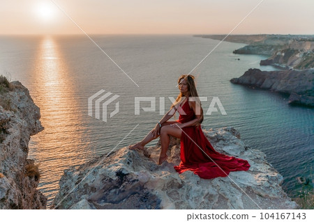 Woman sunset sea red dress, side view a happy beautiful sensual woman in a red long dress posing on a rock high above the sea on sunset. Woman sunset sea red dress, side view a happy beautiful sensual woman in a red long dress posing on a rock high above the sea on sunset. 104167143