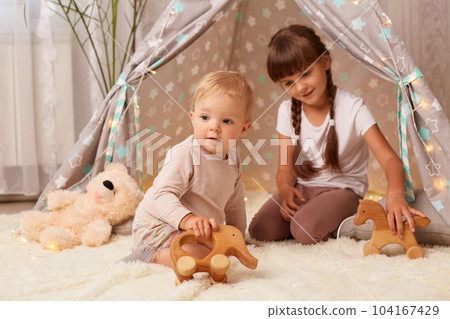 Horizontal shot of positive optimistic sibling enjoying time at home, little girl with braids playing with wooden toys in the wigwam together with her toddler sister. Horizontal shot of positive optimistic sibling enjoying time at home, little girl with braids playing with wooden toys in the wigwam together with her toddler sister. 104167429