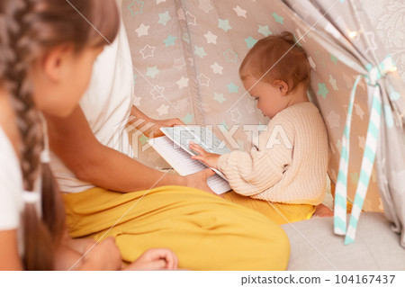 Closeup portrait of infant baby posing in wigwam with her sister with braids and faceless mother sitting near her, charming kid holding book in hands, playing with her family in teepee tent; 104167437