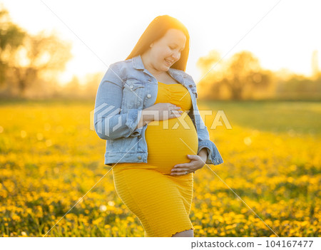 Young pregnant woman smilling to huge belly during lovely summer sunset on the meadow, sun flare, pregnancy concept 104167477