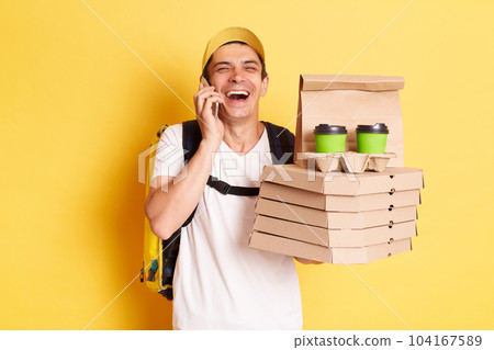 Indoor shot of happy laughing man courier with thermo backpack in white T-shirt and cap isolated over yellow background, talking phone, holding boxes with pizza and takeaway coffee. Indoor shot of happy laughing man courier with thermo backpack in white T-shirt and cap isolated over yellow background, talking phone, holding boxes with pizza and takeaway coffee. 104167589