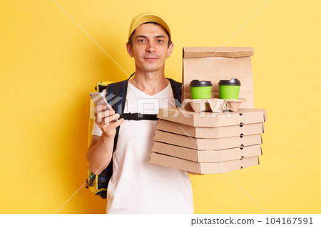Horizontal shot of serious calm man courier with thermo backpack in white T-shirt and cap, looking at camera, holding client's order, standing with smart phone in hands isolated over yellow background 104167591