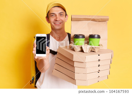 Indoor shot of happy kind man courier with thermo backpack in white T-shirt and cap isolated over yellow background, showing cell phone with empty display for advertisement, holding pizza and coffee. 104167593