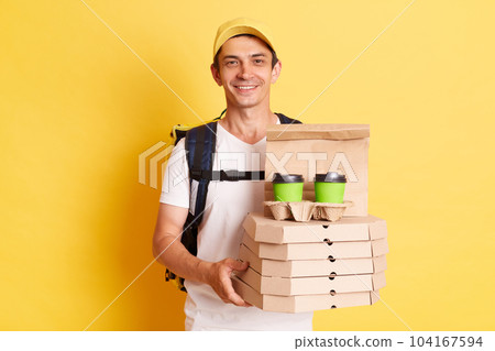 Image of satisfied man courier with thermo backpack in white T-shirt and cap isolated over yellow background, holding boxes with pizza and disposable cups with coffee or tea, delivering order. 104167594