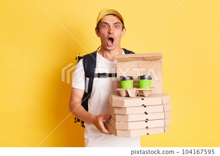 Shocked surprised man courier with thermo backpack in white T-shirt and cap isolated over yellow background, holding boxes with pizza and disposable cups with coffee or tea, delivering order. 104167595
