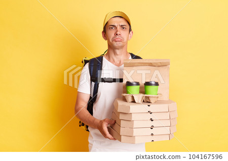 Horizontal shot of sad delivery man wearing white t shirt holding food order pizza boxes and coffee, looking at camera with pout lips and expressing sadness, posing isolated over yellow background. 104167596