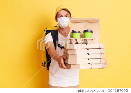 Portrait of delivery employee man in yellow cap, white t-shirt and protective face mask holding cardboard pizza and coffee, handing order to client, isolated on yellow background 104167600