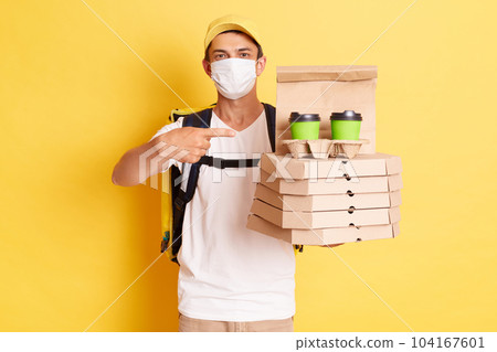 Portrait of deliveryman with thermo backpack in white T-shirt, cap and protective mask standing against yellow wall, pointing at pizza boxes and disposable cups with coffee. Portrait of deliveryman with thermo backpack in white T-shirt, cap and protective mask standing against yellow wall, pointing at pizza boxes and disposable cups with coffee. 104167601