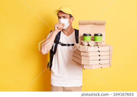 Delivery man wearing protective mask, holding pizza boxes and takeaway coffee, courier working during flu epidemic, catching cold, having influenza symptoms, standing isolated over yellow background. 104167603