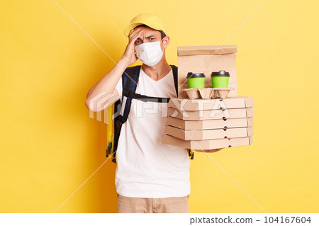 Indoor shot of tired deliveryman in protective mask standing against yellow wall, holding pizza and coffee, suffering headache, forgot clint address, working during quarantine. 104167604