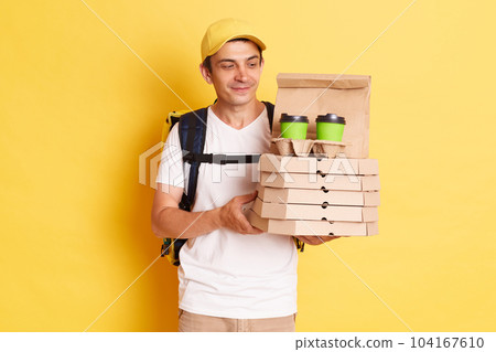 Image of calm smiling deliveryman with thermo backpack in white T-shirt and cap standing against yellow wall, holding and looking at pizza boxes and disposable cups for delivering. Image of calm smiling deliveryman with thermo backpack in white T-shirt and cap standing against yellow wall, holding and looking at pizza boxes and disposable cups for delivering. 104167610