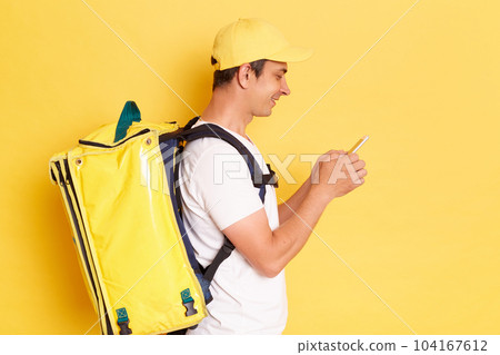 Side view portrait of positive handsome deliveryman with thermo backpack in white T-shirt and cap standing against yellow wall, standing using cell phone, chatting with client. Side view portrait of positive handsome deliveryman with thermo backpack in white T-shirt and cap standing against yellow wall, standing using cell phone, chatting with client. 104167612