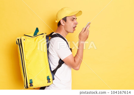 Side view of aggressive courier man with thermo backpack on his back, delivering fast food, holding smart phone and screaming with anger, talking to client or boss, isolated over yellow background. Side view of aggressive courier man with thermo backpack on his back, delivering fast food, holding smart phone and screaming with anger, talking to client or boss, isolated over yellow background. 104167614