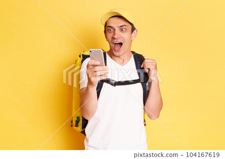 Portrait of excited happy deliveryman with thermo backpack in white T-shirt and cap holding mobile phone in hands, expressing happiness, keeps mouth opened, standing against yellow wall. Portrait of excited happy deliveryman with thermo backpack in white T-shirt and cap holding mobile phone in hands, expressing happiness, keeps mouth opened, standing against yellow wall. 104167619