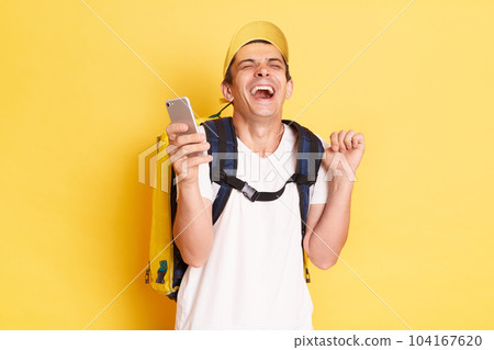 Extremely happy funny delivery man with thermo bag holding mobile phone, using cell phone and laughing, having fun during his work, standing isolated on yellow background. 104167620