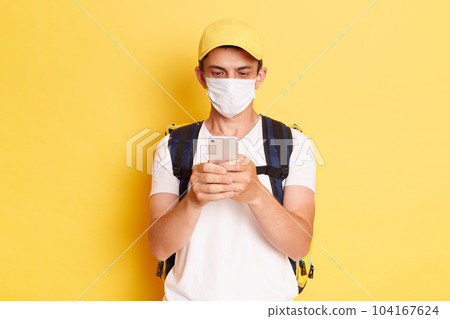 indoor shot of delivery man in yellow cap, t-shirt and protective mask, with thermal food backpack using cell phone while standing isolated on yellow background. indoor shot of delivery man in yellow cap, t-shirt and protective mask, with thermal food backpack using cell phone while standing isolated on yellow background. 104167624