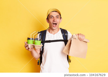 Portrait of astonished deliveryman wearing white t shirt and cap holding paper package and take away coffee in hands, fast delivery, courier posing isolated on yellow background. 104167629