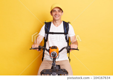 Food delivery. Portrait of smiling positive courier guy riding bike with backpack bag delivering meal from takeaway cafe or restaurant isolated over yellow background, Food delivery. Portrait of smiling positive courier guy riding bike with backpack bag delivering meal from takeaway cafe or restaurant isolated over yellow background, 104167634