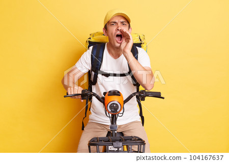 Indoor shot of excited deliveryman on bicycle wearing white T-shirt and cap isolated over yellow background, screaming announcement loud, keeps hand near mouth, delivering orders. Indoor shot of excited deliveryman on bicycle wearing white T-shirt and cap isolated over yellow background, screaming announcement loud, keeps hand near mouth, delivering orders. 104167637