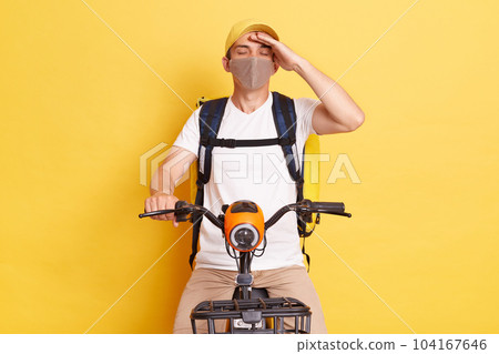 Tired exhausted delivery man in protective mask with thermo backpack on electric scooter, having terrible headache, has flu symptoms, posing with closed eyes isolated on yellow background. Tired exhausted delivery man in protective mask with thermo backpack on electric scooter, having terrible headache, has flu symptoms, posing with closed eyes isolated on yellow background. 104167646