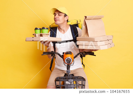 Satisfied deliveryman on bicycle holding paper parcel, pizza boxes and disposable coffee cups, smelling hot beverage, enjoying aroma while delivering food, isolated over yellow background. Satisfied deliveryman on bicycle holding paper parcel, pizza boxes and disposable coffee cups, smelling hot beverage, enjoying aroma while delivering food, isolated over yellow background. 104167655