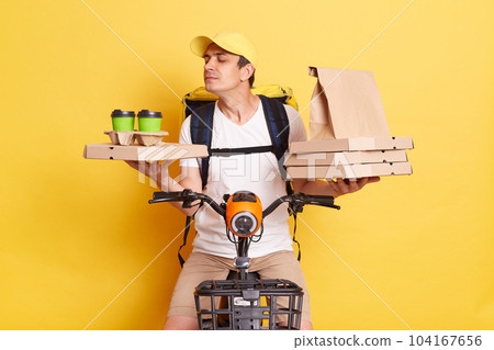 Horizontal shot of courier wearing cap and white t shirt holding pizza boxes and takeaway drink, riding bicycle, posing isolated on yellow background, smelling and enjoying tasty coffee. 104167656