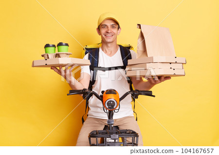 Positive courier man on bike being in hurry to delivery food to clint, holding carton pizza boxes and disposable cups with hot beverage, wearing white t shirt and cap, isolated over yellow background. 104167657