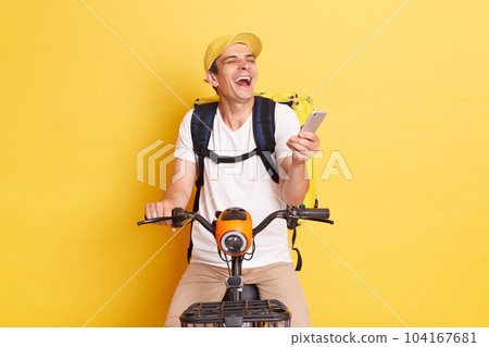 Indoor shot of happy young delivery guy holding cell phone, wearing cap and thermo bag, riding bicycle, laughing, expressing positive emotions, isolated on yellow background Indoor shot of happy young delivery guy holding cell phone, wearing cap and thermo bag, riding bicycle, laughing, expressing positive emotions, isolated on yellow background 104167681