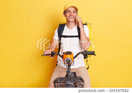 Horizontal shot of smiling delighted pleased courier man holding pizza in carton boxes, wearing white T-shirt, riding electric scooter, posing against yellow wall, talking phone with client. 104167682