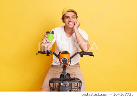 Image of satisfied man wearing white t shirt and cap riding electric scooter and stops to drink takeaway coffee, enjoying his weekend, isolated on yellow background. 104167723