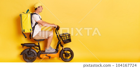 Side view portrait of delivery man with backpack in white T-shirt and cap riding e scooter, being scared of accident, looks scared and shocked, isolated over yellow background. Side view portrait of delivery man with backpack in white T-shirt and cap riding e scooter, being scared of accident, looks scared and shocked, isolated over yellow background. 104167724