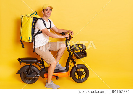Full length profile portrait of man courier with thermo backpack in white T-shirt and cap isolated over yellow background, riding electric bike, delivering orders to clients. 104167725