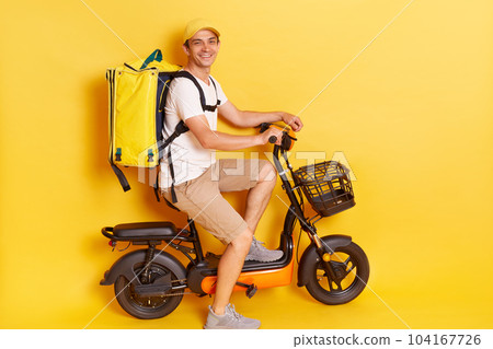 Delivery man wearing cap and t-shirt driving moped motorbike scooter, posing isolated on yellow background, guy employee working as courier, looking at camera with positive expression. 104167726