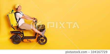 Full length side view portrait of man courier with thermo backpack in white T-shirt and cap riding electric bike, braking, avoids accident, looks scared and shocked, isolated over yellow background. 104167729