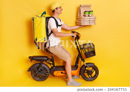 Profile portrait of courier man wearing cap and t-shirt driving electric bike motorbike handing pizza in cardboard and takeaway coffee isolated on yellow background. Profile portrait of courier man wearing cap and t-shirt driving electric bike motorbike handing pizza in cardboard and takeaway coffee isolated on yellow background. 104167731