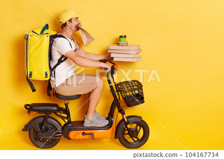 Profile portrait of young man with backpack wearing white t shirt riding e-scooter with pizza boxes and coffee, holding paper parcel in teeth, delivering lots orders, isolated over yellow background. 104167734