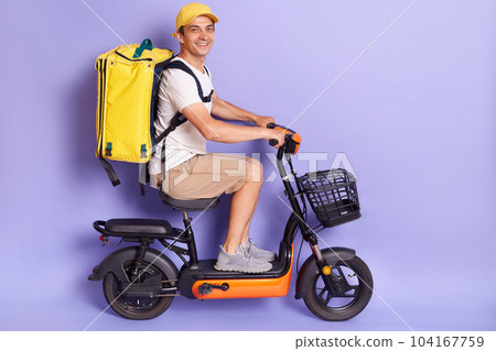 Indoor shot of delivery man wearing with thermo backpack delivering food on electric scooter isolated over purple background, riding to client with fast food order, looking at camera. Indoor shot of delivery man wearing with thermo backpack delivering food on electric scooter isolated over purple background, riding to client with fast food order, looking at camera. 104167759
