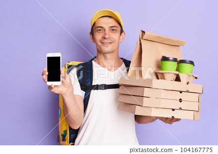 Photo of smiling attractive man courier in yellow cap and T-shirt showing food order and smart phone with empty display, bringing pizza boxes and coffee to go to client, isolated on purple background. Photo of smiling attractive man courier in yellow cap and T-shirt showing food order and smart phone with empty display, bringing pizza boxes and coffee to go to client, isolated on purple background. 104167775