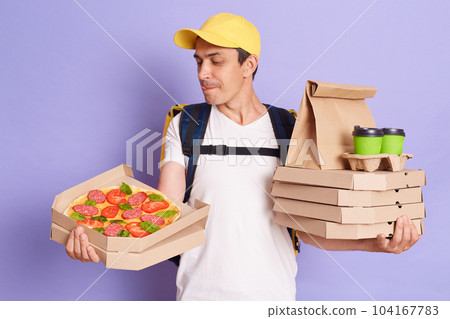 Portrait of hungry man courier holds pizza boxes in hand isolated on purple background, looking at tasty fast food with sticking tongue, wants to eat while delivering orders. Portrait of hungry man courier holds pizza boxes in hand isolated on purple background, looking at tasty fast food with sticking tongue, wants to eat while delivering orders. 104167783