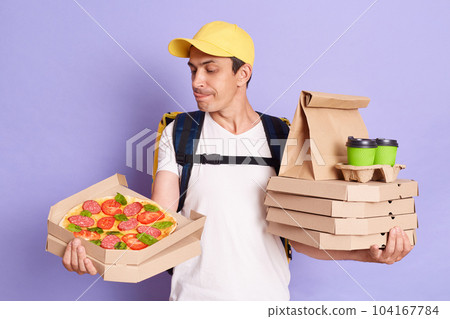 Indoor shot of young adult man courier wearing yellow cap holding pizza boxes, takeaway coffee and showing tongue out, feels hungry, wants to eat somebody order. Indoor shot of young adult man courier wearing yellow cap holding pizza boxes, takeaway coffee and showing tongue out, feels hungry, wants to eat somebody order. 104167784