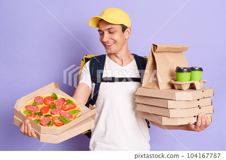 Indoor shot of smiling delivery man wearing white T-shirt with thermo backpack holding pizza boxes and coffee in disposable cups isolated over purple background, showing order to client. 104167787