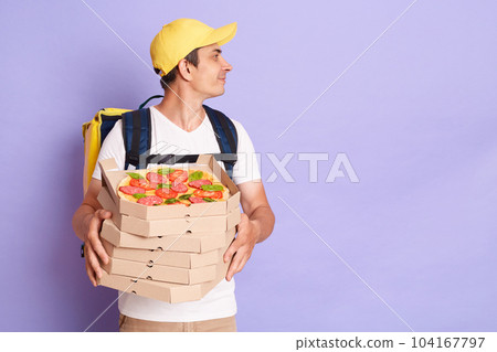 Portrait of delivery man wearing white T-shirt with thermo backpack holding pizza boxes isolated over purple background, looking away at advertisement area, copy space for promotion. 104167797