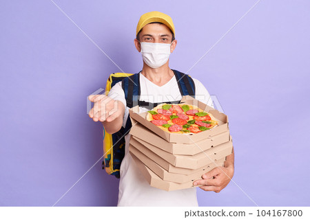 Delivery man in cap, t-shirt and mask giving food order pizza boxes isolated on lilac background, stretching hand to client, waiting his money. Service during coronavirus pandemic and quarantine. Delivery man in cap, t-shirt and mask giving food order pizza boxes isolated on lilac background, stretching hand to client, waiting his money. Service during coronavirus pandemic and quarantine. 104167800