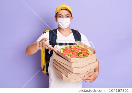 Portrait of courier man wearing white T-shirt and protective with thermo backpack holding pizza boxes, delivering food during quarantine, posing isolated over purple background. Portrait of courier man wearing white T-shirt and protective with thermo backpack holding pizza boxes, delivering food during quarantine, posing isolated over purple background. 104167801